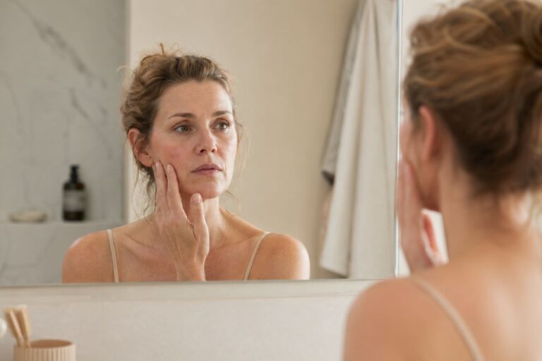 empfindliche Haut ab 30 pflegen – Frau um die 40 betrachtet nachdenklich ihre Haut im Spiegel und berührt ihr Gesicht, minimalistische Badezimmerszene