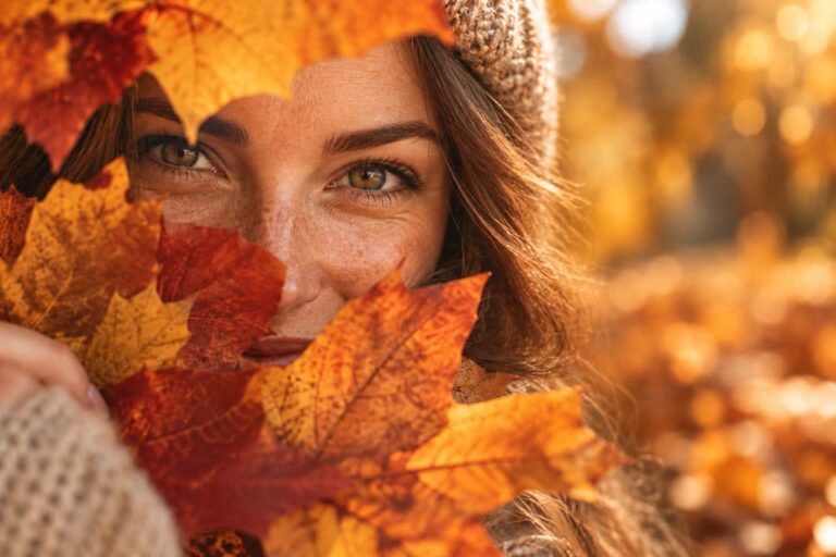 Hautpflege im Herbst – Frau mit Strickmütze blickt durch buntes Herbstlaub – Symbolbild Hautpflege im Herbst